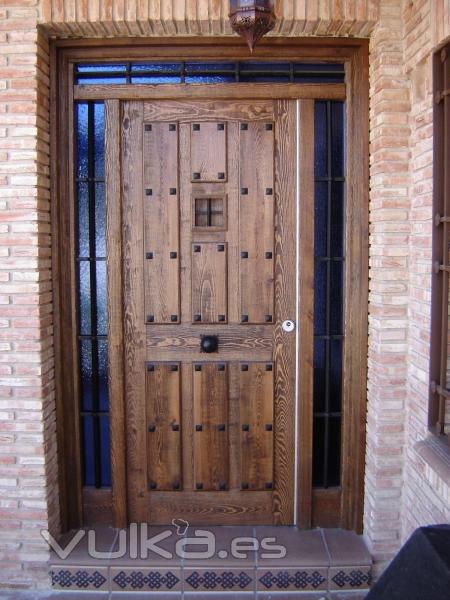 Puerta de entrada en Pino teñida en Nogal, modelo tablas tipo posada, con dos muros y montante fijos, reja de ...