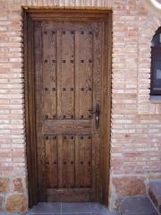 Puerta de entrada en pino, teida en nogal, modelo tablas tipo posada con herrajes negros.